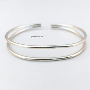 Può includere: Un bracciale a fascia in metallo color argento con un design a doppio cerchio e un'apertura sulla parte superiore. Il testo 925 SilverThread è stampato in nero al centro dello spazio aperto tra le due fasce metalliche lisce e lucide.