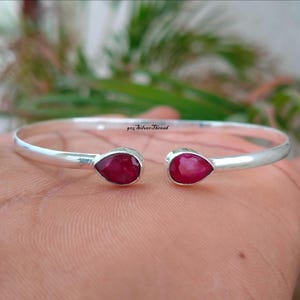 Op de afbeelding: Een zilveren open manchetarmband met twee druppelvormige, dieprode edelstenen. De armband heeft een eenvoudig ontwerp, met de edelstenen aan elk uiteinde. De tekst "925 SilverThread" is zichtbaar.