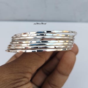 Op de afbeelding: Een set van zeven gestapelde zilveren armbanden met een gehamerd, getextureerd oppervlak. Elke armband is gemaakt van gepolijst sterling zilver, wat zorgt voor een reflecterende, metalen uitstraling. De tekst 925 SilverThread is zichtbaar.