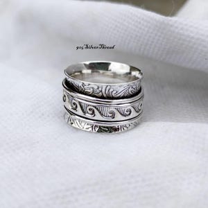 Könnte beinhalten: Ein Spinner-Ring aus Sterlingsilber mit drei verschiedenen Bändern. Die oberen und unteren Bänder zeigen komplizierte florale Schnörkel, während das mittlere Band ein sich wiederholendes Wellenmuster aufweist. Text 925 SilverThread sichtbar.