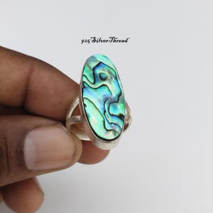 May include: An oval-shaped ring featuring a polished abalone shell inlay with iridescent green, blue, and black swirls set in a silver band. The text 925 SilverThread is visible above the ring. The design showcases a smooth, metallic finish.
