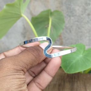 Pode incluir: Uma pulseira rígida de tom prateado polido com uma banda de metal lisa e contínua que se curva numa forma de onda distinta no centro. A pulseira tem um acabamento metálico refletor e um design simples e aberto.