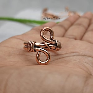 Può includere: Un anello in filo di rame con design bypass, due anelli aperti alle estremità e un avvolgimento decorativo in filo attorno alla fascia. Il metallo ha una finitura lucida color marrone rossastro caldo. Il testo 925 SilverThread è visibile.