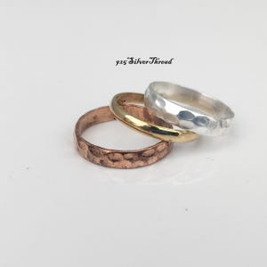 Puede incluir: Tres anillos apilados que incluyen un anillo de cobre con textura martillada, un anillo liso de tono dorado y un anillo de plata con acabado martillado. El texto 925 SilverThread es visible sobre los anillos. Estos aros metálicos están agrupados.