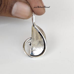 Puede incluir: Un colgante de plata de ley con una placa metálica pulida, curva y semicircular, enmarcada por un fino bucle de hilo de plata con un pequeño acento esférico. Incluye una anilla superior para colgar y el texto 925 SilverThread.