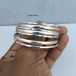 Könnte beinhalten: Ein Stapel aus fünf silbernen Armreifen mit einer gehämmerten, strukturierten Oberfläche. Die Armbänder sind metallisch und reflektierend und haben ein abgerundetes Profil. Der Text 925 SilverThread ist im oberen Bildbereich sichtbar.