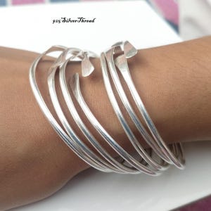 Peut inclure: Un bracelet jonc en argent à plusieurs rangs avec une finition lisse et polie et des extrémités effilées. Le design se compose de plusieurs boucles interconnectées qui s'enroulent autour du poignet, créant un aspect superposé. Le texte 925 SilverThread est visible.