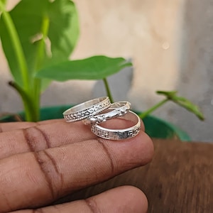 May include: A set of three silver-toned metal rings stacked together. The top ring features a beaded texture, the middle ring has a series of small raised oval shapes, and the bottom ring displays a repeating geometric pattern of small square symbols.