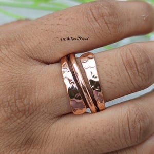 May include: A set of three copper-colored rings featuring a hammered, textured finish. The rings are stacked together, with the central band appearing thinner and smoother than the two outer bands. The text 925 SilverThread is visible.