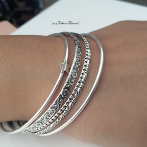 May include: A set of four silver bangles featuring a smooth band with a small star charm, a floral engraved band, a textured geometric band, and a plain polished band. The text 925 SilverThread is visible above the bracelets on the white surface.