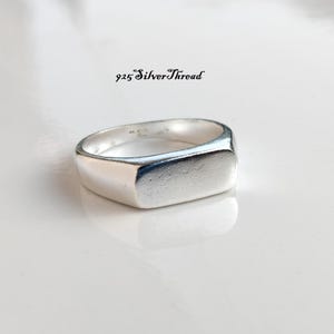Peut inclure: Une bague chevalière en argent poli avec une face rectangulaire plate et un anneau lisse. Le texte 925 SilverThread est imprimé en noir au-dessus de la bague. La bague présente un éclat métallique et une structure solide réfléchissant la lumière.