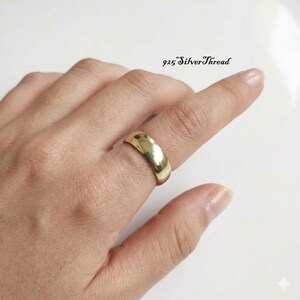 Puede incluir: Un anillo de banda liso y pulido en tono dorado con un perfil redondeado. El anillo se lleva en un dedo y presenta el texto 925 SilverThread impreso en negro en la esquina superior derecha del marco, indicando el nombre de la marca.