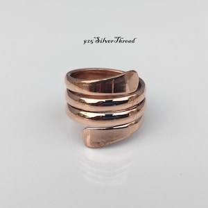 May include: A rose gold-toned metal ring featuring a multi-coil wrap design with smooth, rounded edges. The text 925 SilverThread is printed in black above the ring. The band spirals around itself, creating a layered appearance with a polished finish.