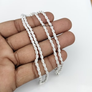 Può includere: Una collana a catena a corda in argento brillante con un design a maglie metalliche intrecciate. La catena ha una finitura lucida e riflettente e include una piccola etichetta con il testo 925 SilverThread, che indica la composizione del gioiello.