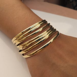 May include: A set of six polished gold-tone metal bangles worn together. Each bracelet features a smooth, rounded, and reflective surface with a consistent circular shape, creating a stacked appearance on the wrist with uniform metallic luster.