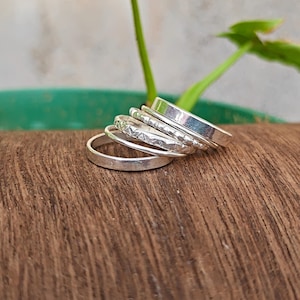 May include: A stack of five silver-colored rings resting on a wooden surface. The set includes one smooth band, one band with a textured, hammered finish, and three additional plain bands of varying widths, all arranged in a cascading, overlapping formation.
