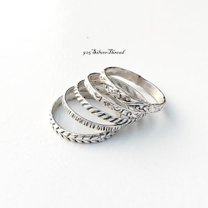 Puede incluir: Un conjunto de cinco anillos de plata con texturas y patrones variados, incluyendo motivos de hojas, diseños de cuerda trenzada y grabados florales. El texto 925 SilverThread es visible sobre los anillos dispuestos en cascada.