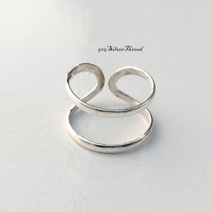 May include: A silver ring featuring a double band design with an open top, where the upper band curves into two circular loops. The text 925 SilverThread is printed in black above the ring, indicating the material composition of the jewellery.