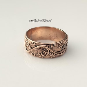 Op de afbeelding: Een koperkleurige metalen ring met een reliëfpatroon van bloemen en ranken op de buitenste band. De tekst 925 SilverThread is in het zwart boven de ring gedrukt. De band heeft een textuurafwerking met verhoogde details van kleine bloemen.