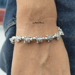 Puede incluir: Una pulsera de plata que presenta una serie de pequeños y detallados dijes de tortuga unidos por diminutas cuentas de plata. El metal tiene un acabado oxidado para resaltar los patrones de los caparazones. El texto 925 Silver Thread es visible.