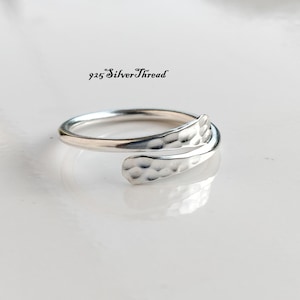 Puede incluir: Un anillo de plata tipo bypass con textura martillada en los extremos superpuestos. El anillo lleva el texto 925 SilverThread. La banda tiene un acabado pulido y un diseño abierto que permite ajustarlo a diferentes tamaños de dedo.