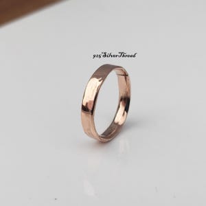 Pode incluir: Um anel de metal em tom de ouro rosa com uma superfície lisa e polida e um perfil de banda plano. O anel está posicionado na vertical, exibindo um acabamento reflexivo. O texto 925 SilverThread está impresso a preto acima do anel.