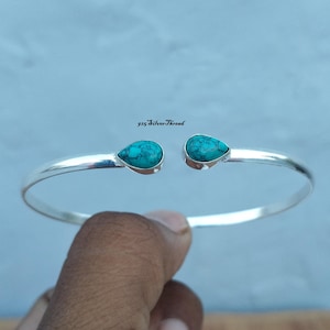 May include: A silver cuff bracelet featuring two teardrop-shaped turquoise stones with dark veining at the open ends. The metal band has a polished finish. The text 925 SilverThread is visible above the centre gap of the bracelet.
