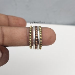 Könnte beinhalten: Ein stapelbares Ring-Set bestehend aus fünf einzelnen Ringen in abwechselnden Gold-, Silber- und Kupfertönen. Die Ringe weisen unterschiedliche Texturen auf, darunter glatte Oberflächen und Perlenverzierungen. Der Text 925 SilverThread ist sichtbar.