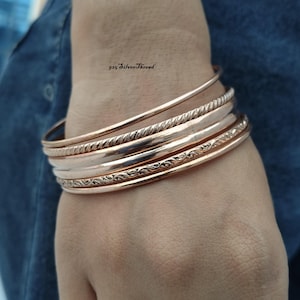 Peut inclure: Un ensemble de cinq bracelets joncs couleur or rose présentant des textures variées, notamment des motifs lisses, torsadés et gravés. Le texte 925 SilverThread est visible au-dessus des bracelets. Ces joncs métalliques sont portés ensemble.