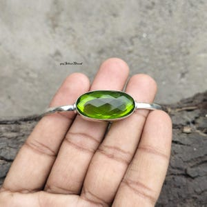 Op de afbeelding: Een zilveren armband met een grote, ovaalvormige, gefacetteerde peridotgroene edelsteen in het midden. De metalen band is glad en gepolijst en loopt door aan beide zijden van de zetting. De tekst 925 Silver Thread is zichtbaar.