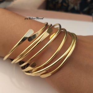 Può includere: Un set di diversi bracciali a fascia in metallo color oro con una finitura liscia e lucida. I bracciali presentano un design aperto e avvolgente con estremità arrotondate. Il testo 925 SilverThread è visibile nella parte superiore.