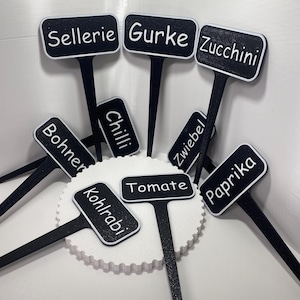 Könnte beinhalten: Eine Sammlung schwarzer Gartenstecker mit weißen Texten. Die Etiketten tragen Namen wie "Zucchini", "Tomate" und "Chili". Jeder Stecker hat ein rechteckiges Etikett mit weißem Rand und einen langen, dünnen Stiel zum Einsetzen in die Erde. Die Stecker sind auf einer weißen, strukturierten Oberfläche angeordnet.
