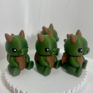 Könnte beinhalten: Vier grüne Drachenfiguren mit braunen Akzenten, darunter Hörner, Flügel und Bauch. Die Drachen haben große schwarze Augen und sitzen auf einer weißen, kreisförmigen Plattform. Die Figuren sind wahrscheinlich handgefertigt und für Fantasy-Dekoration geeignet.