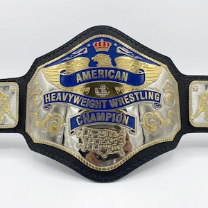 May include: A championship wrestling belt featuring a black leather strap and a large central metal plate. The plate displays a gold eagle, a map of the United States, and blue banners with the text AMERICAN HEAVYWEIGHT WRESTLING CHAMPION.