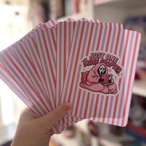 May include: A set of rectangular cards featuring a pink and white vertical striped pattern. The front card displays a graphic of a ghost character wrapped in a pink blanket, reading a book, with the text "JUST ONE MORE CHAPTER" above it.