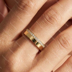 Könnte beinhalten: Ein goldener Bandring mit einer Reihe kleiner, klarer Edelsteine ​​am oberen Rand. Der Ring befindet sich an einem Finger, wobei heller Hautton sichtbar ist. Der Ring scheint neu und ungetragen zu sein.