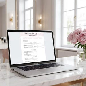 May include: A laptop displaying a document with the name "Valentina Liddi" sits on a marble desk. Makeup brushes, perfume, and a vase of pink peonies are also on the desk. The background features a vanity and a window.
