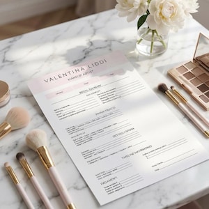 Può includere: Un'area di lavoro di una truccatrice con un modulo di riepilogo nuziale, pennelli per il trucco e una palette di ombretti. Il modulo è intitolato "VALENTINA LIDDI MAKEUP ARTIST". Peonie bianche in un vaso di vetro aggiungono un tocco di eleganza. I pennelli hanno manici rosa e dettagli dorati.