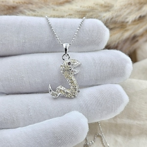 Op de afbeelding: Een zilverkleurige slangenketting met een hanger in de vorm van een kronkelende slang, ingelegd met talloze kleine, heldere en fonkelende strass-steentjes. De hanger hangt aan een fijne zilverkleurige bolletjesketting voor een glinsterend effect.