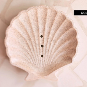 May include: A light beige seashell-shaped soap dish with a textured surface and three drainage holes. The stone dish has the text "OCEAN STONE" in a black rectangle. The dish is made of stone.