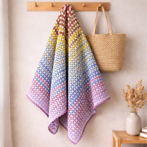 Puede incluir: Una colorida manta de ganchillo con un patrón de cuadrados de la abuela cuelga de un perchero de madera. La manta presenta un degradado de colores, incluyendo rojo, amarillo, azul y morado. También se ve un bolso tote tejido y un jarrón con flores secas.
