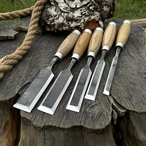 Puede incluir: Un juego de cinco cinceles de madera con mangos de madera y férulas de metal plateado. Los cinceles están dispuestos sobre una superficie de madera envejecida, con una funda de cuero visible en el fondo. Una gruesa cuerda está sobre la madera.
