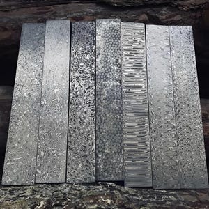 May include: A collection of eight rectangular metal pieces with intricate, swirling patterns in shades of gray and silver. Each piece displays a unique design, showcasing a variety of textures and visual effects. The pieces are arranged vertically, side by side.