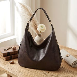 Puede incluir: Un bolso hobo de cuero marr&oacute;n oscuro con una sola correa para el hombro. El bolso tiene forma curva, cierre de cremallera y un peque&ntilde;o detalle de borla. El bolso est&aacute; sobre una superficie de madera.