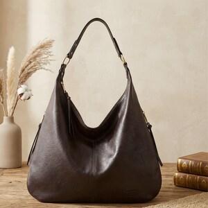 Puede incluir: Un bolso hobo de cuero marr&oacute;n oscuro con una sola correa para el hombro. El bolso tiene forma curva y cierre de cremallera con borla. El bolso est&aacute; sobre una superficie de madera junto a un jarr&oacute;n con flores secas y libros.