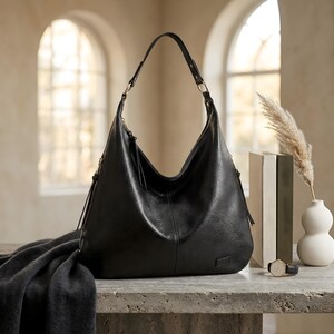 Puede incluir: Un bolso hobo grande de cuero negro con una sola correa para el hombro. El bolso tiene forma curva y cierre de cremallera. Una bufanda gris oscuro est&aacute; sobre la mesa frente al bolso. Un reloj dorado y un jarr&oacute;n blanco tambi&eacute;n est&aacute;n sobre la mesa.