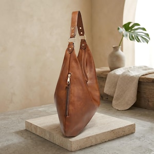 Puede incluir: Un bolso hobo de cuero marr&oacute;n con una sola correa para el hombro. El bolso tiene un bolsillo con cremallera en la parte delantera y herrajes dorados. El bolso est&aacute; sobre una superficie de piedra.