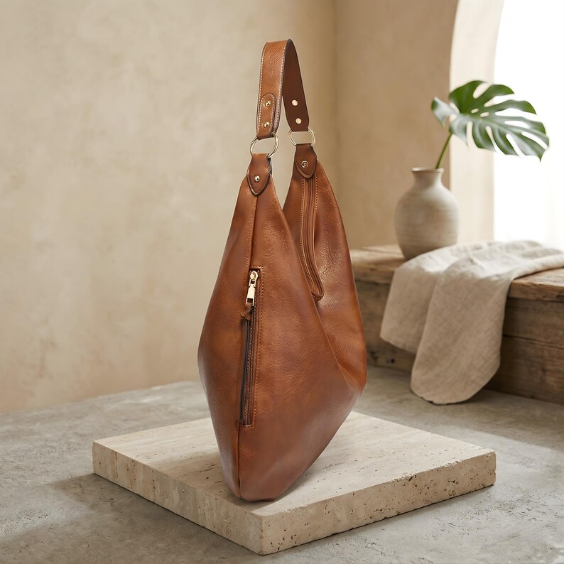 Puede incluir: Un bolso hobo de cuero marr&oacute;n con una sola correa para el hombro. El bolso tiene un bolsillo con cremallera en la parte delantera y herrajes dorados. El bolso est&aacute; sobre una superficie de piedra.