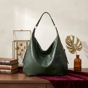 Puede incluir: Bolso de hombro de cuero verde oscuro con una sola correa y un dise&ntilde;o curvo. El bolso tiene una correa larga y una borla a cada lado. El bolso est&aacute; sobre una superficie de madera con un pa&ntilde;o de terciopelo rojo.