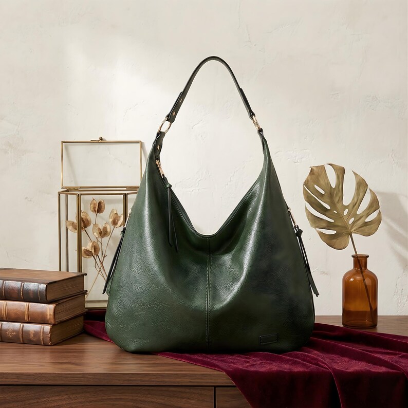 Puede incluir: Bolso de hombro de cuero verde oscuro con una sola correa y un dise&ntilde;o curvo. El bolso tiene una correa larga y una borla a cada lado. El bolso est&aacute; sobre una superficie de madera con un pa&ntilde;o de terciopelo rojo.